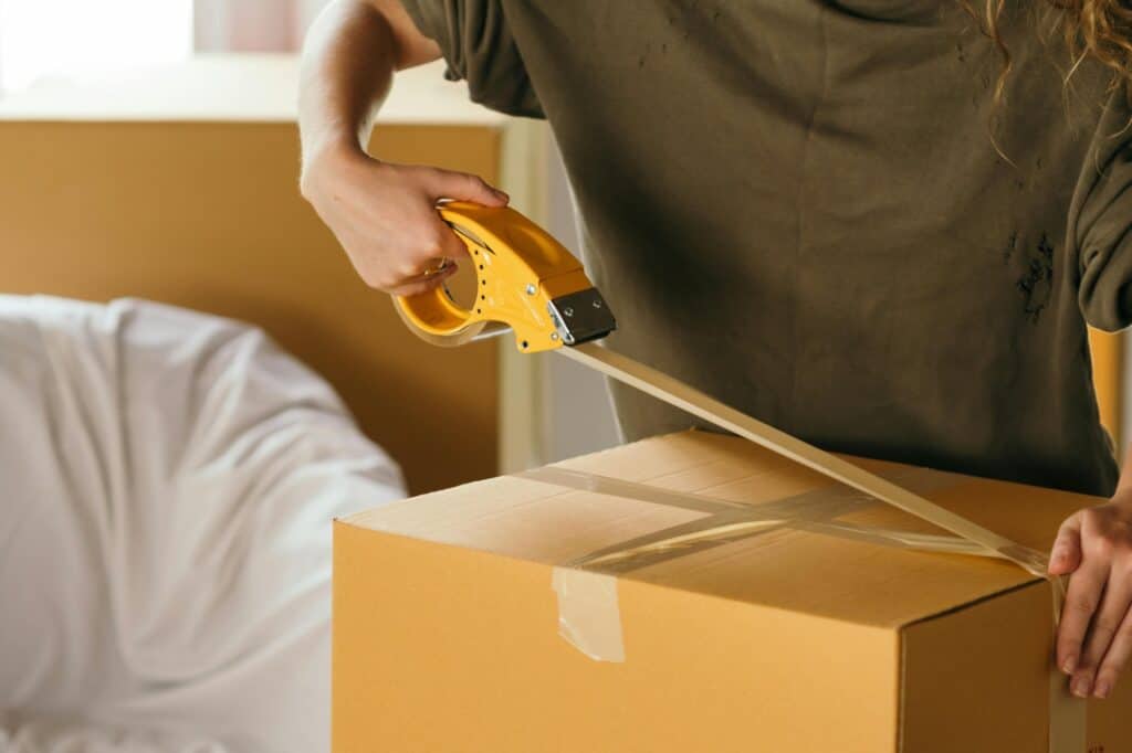 A woman is sealing a carton while a wrapped sofa is on the background; in the concept of moving to Sutton.